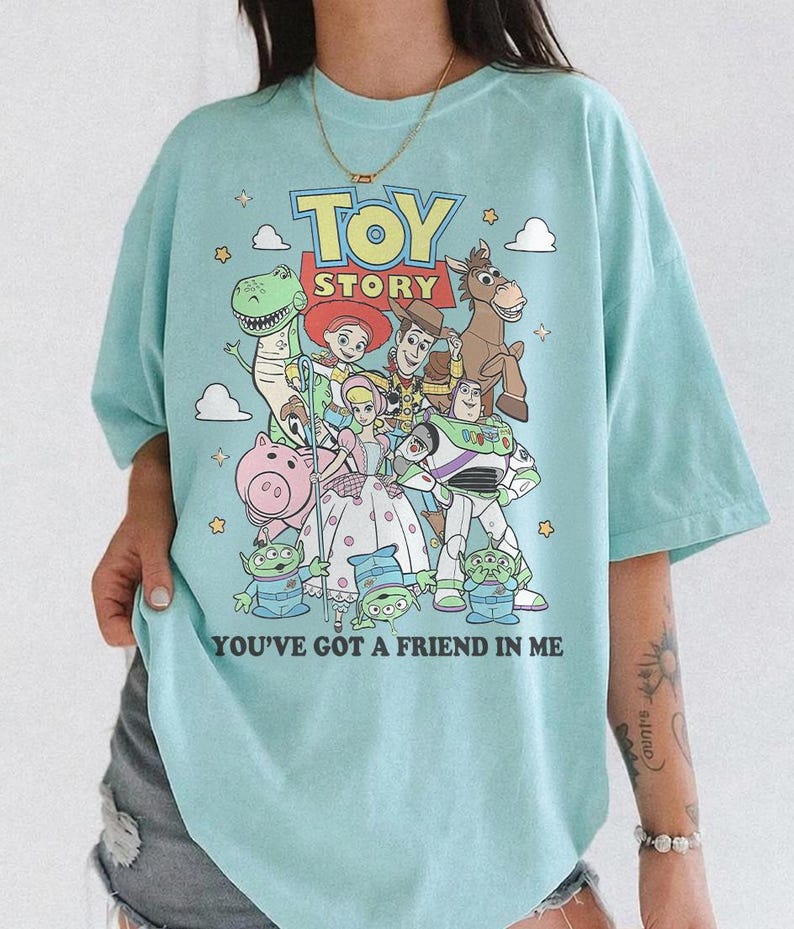 May include: A light blue t-shirt featuring a colorful graphic print of Toy Story characters including Woody, Buzz Lightyear, Jessie, Rex, Bullseye, Bo Peep, Hamm, and the Aliens, with the text TOY STORY and YOU'VE GOT A FRIEND IN ME printed on the front.
