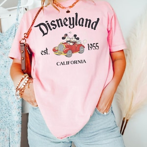 K&ouml;nnte beinhalten: Rosa T-Shirt mit Disneyland-Grafik. Die Grafik zeigt Micky und Minnie Maus in einem roten Auto mit dem Text "Disneyland est. 1955 CALIFORNIA". Das Shirt hat einen Rundhalsausschnitt und kurze &Auml;rmel.