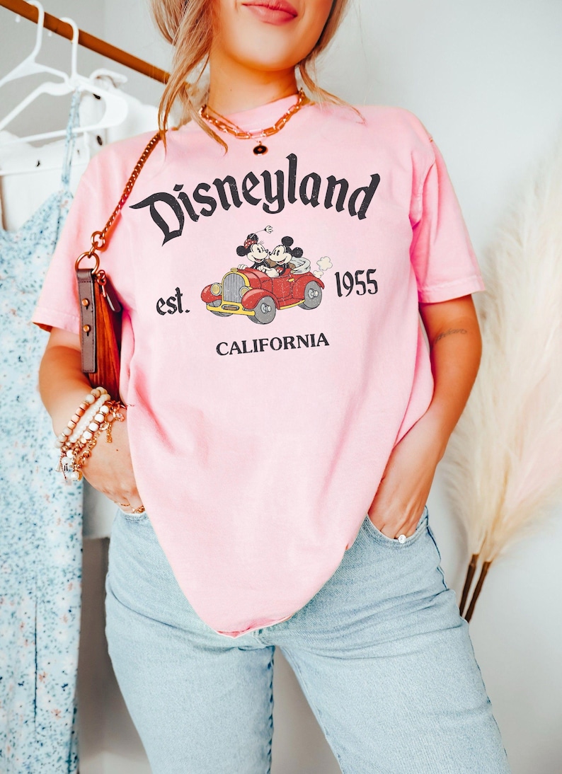 K&ouml;nnte beinhalten: Rosa T-Shirt mit Disneyland-Grafik. Die Grafik zeigt Micky und Minnie Maus in einem roten Auto mit dem Text "Disneyland est. 1955 CALIFORNIA". Das Shirt hat einen Rundhalsausschnitt und kurze &Auml;rmel.