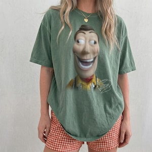May include: A sage green short-sleeve t-shirt featuring a large, centered graphic of a smiling cowboy character with wide eyes. The graphic includes the handwritten-style text "Woody" in white script located to the right of the character's torso.