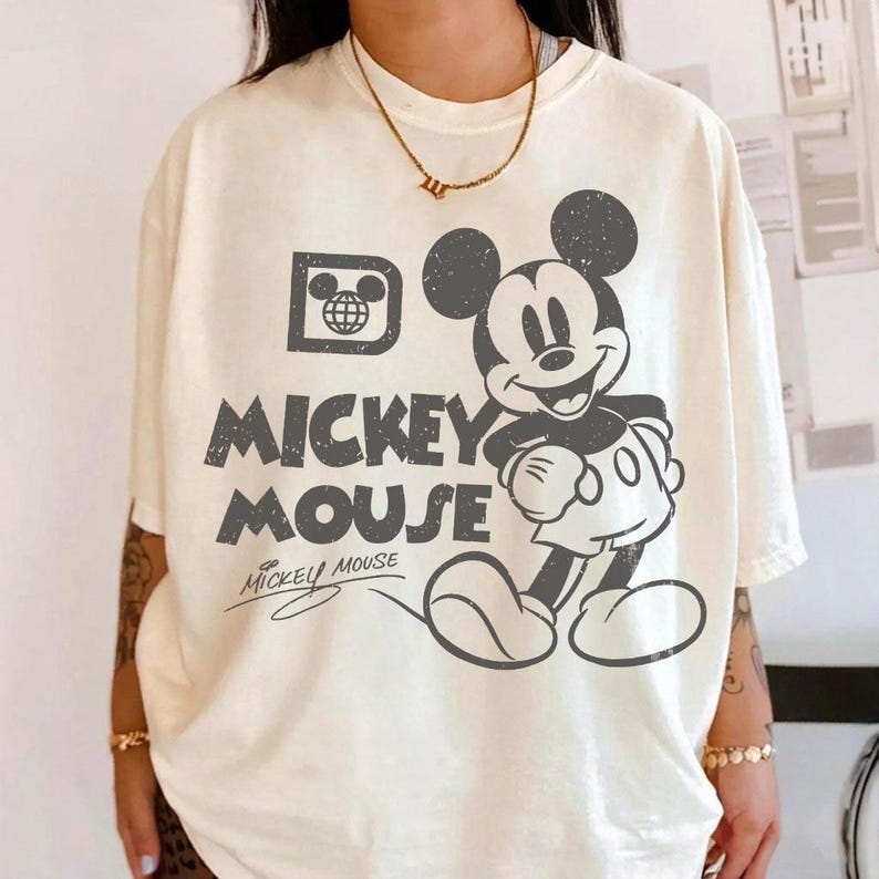 May include: Cream-colored t-shirt featuring a distressed graphic of Mickey Mouse. The design includes the words "Mickey Mouse" in a bold, vintage font, along with a stylized image of Mickey. A classic Disney-themed apparel item.
