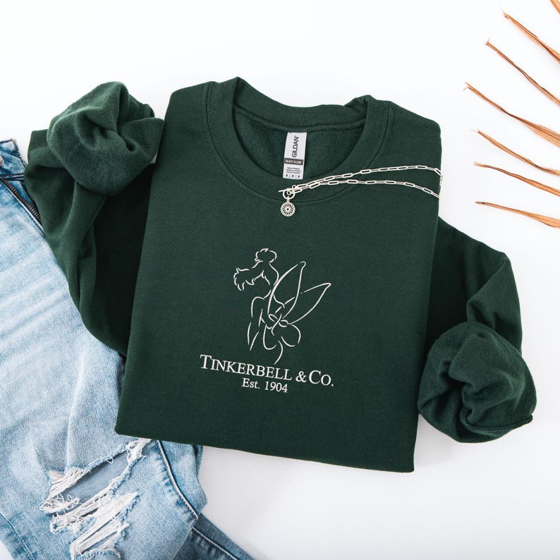 Pode incluir: Uma camisola verde escuro com um gr&aacute;fico branco de uma fada e o texto "Tinkerbell & Co. Est. 1904." Um colar de prata com um pingente est&aacute; sobre a camisola. A camisola est&aacute; ao lado de um par de jeans azuis.