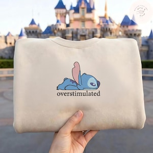 May include: A beige crewneck sweatshirt featuring an embroidered graphic of a blue alien character lying flat on its stomach with one ear raised. Below the character, the word overstimulated is printed in a simple, dark, lowercase font.