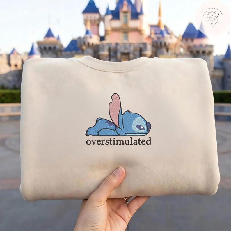 May include: A beige crewneck sweatshirt featuring an embroidered graphic of a blue alien character lying flat on its stomach with one ear raised. Below the character, the word overstimulated is printed in a simple, dark, lowercase font.