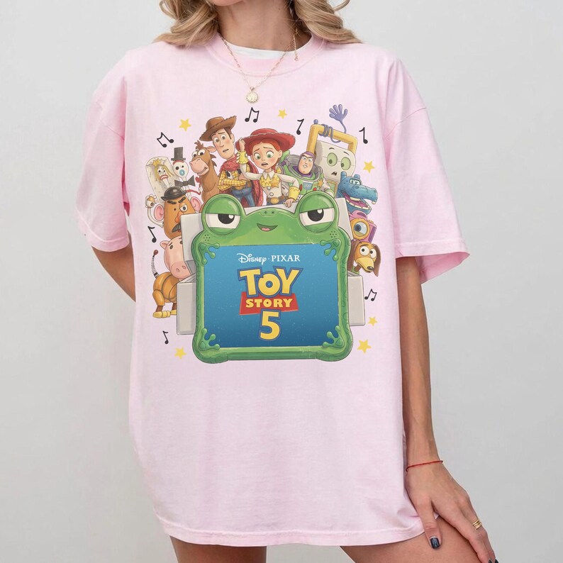Może przedstawiać: Jasnor&oacute;żowy t-shirt z nadrukiem graficznym przedstawiającym r&oacute;żne postacie z Toy Story otaczające zieloną ramkę w kształcie żaby. Wewnątrz ramki znajduje się napis Disney Pixar Toy Story 5. Wz&oacute;r zawiera małe ż&oacute;łte gwiazdki i nuty muzyczne.