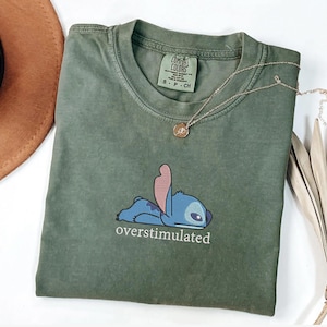 May include: An olive green short-sleeve t-shirt featuring a graphic of the blue character Stitch lying down with the text "overstimulated" printed underneath in white. The shirt has a crew neckline and a Comfort Colors brand label inside.