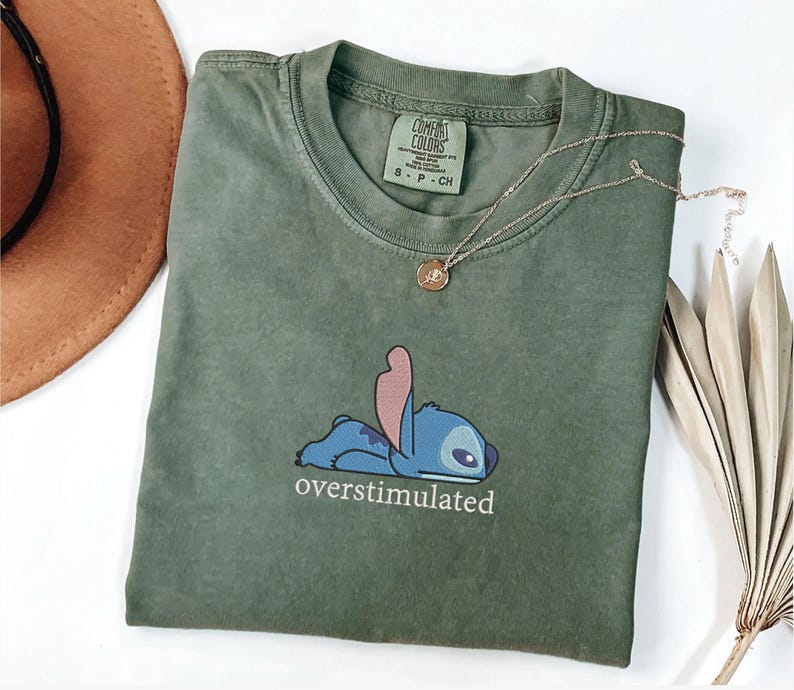 May include: An olive green short-sleeve t-shirt featuring a graphic of the blue character Stitch lying down with the text "overstimulated" printed underneath in white. The shirt has a crew neckline and a Comfort Colors brand label inside.