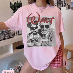 May include: A light pink t-shirt featuring a vintage-style graphic of Mickey Mouse and Walt Disney, with the word "Walt" in red script. The shirt is a soft, comfortable material.