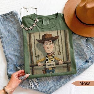 May include: An olive green t-shirt featuring a graphic of the character Woody from Toy Story, wearing a cowboy hat and a sheriff badge, with the text WOODY on a sign. The shirt is paired with blue denim jeans, a brown hat, and a floral necklace.