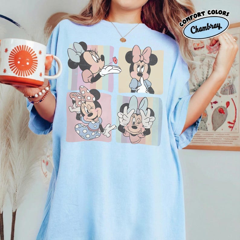 May include: Light blue t-shirt featuring a four-panel graphic of Minnie Mouse in various poses. Each panel has a pastel background. The shirt also has the text "COMFORT COLORS Chambray" in a circular design.