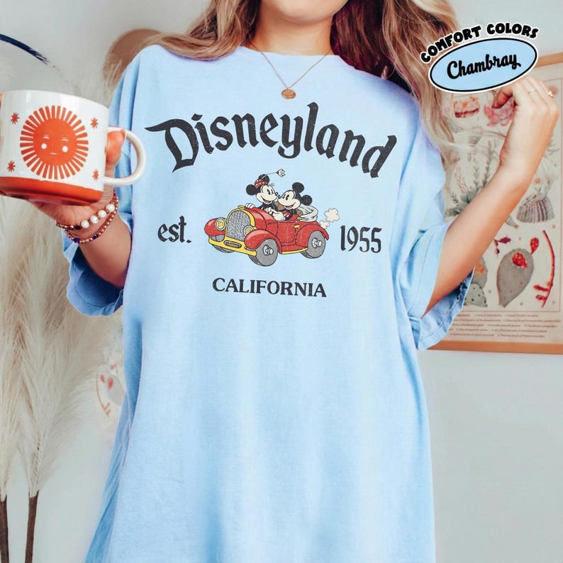 K&ouml;nnte beinhalten: Hellblaues T-Shirt mit dem Disneyland-Logo, das Micky und Minnie Maus in einem roten Auto zeigt. Das Shirt hat den Text "est. 1955 CALIFORNIA". Es ist eine Komfortfarbe und hat ein Chambray-Etikett.