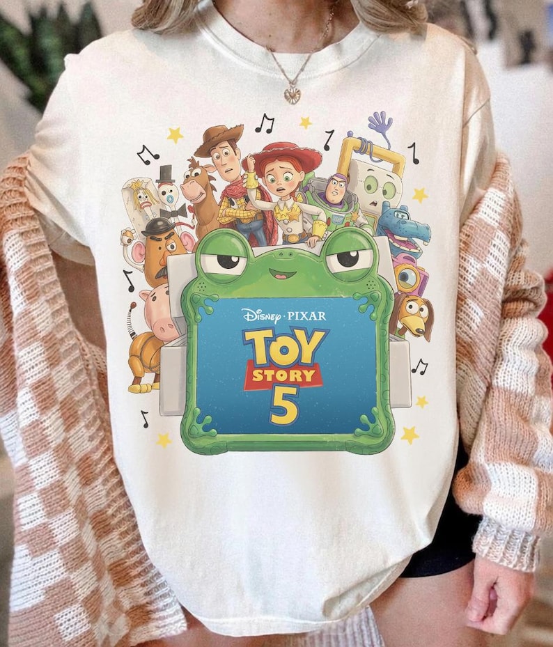 Może przedstawiać: Kremowy t-shirt z nadrukiem graficznym przedstawiającym postacie z filmu Disney Pixar Toy Story, w tym Chudego, Jessie i Buzza Astrala. Duża zielona ramka w kształcie żaby otacza napis Disney Pixar Toy Story 5 z ż&oacute;łtymi gwiazdkami.