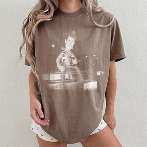 May include: A brown short-sleeve t-shirt featuring a monochromatic graphic print of a cowboy character sitting on the hood of a car. The character wears a hat and vest, holding a drink. The car has a visible license plate with the text 02-882.