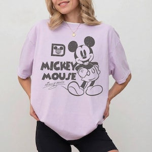 May include: A light purple t-shirt featuring a distressed graphic of Mickey Mouse. The design includes the words "Mickey Mouse" in a bold font, along with Mickey's signature. The shirt has a classic crew neck and short sleeves.