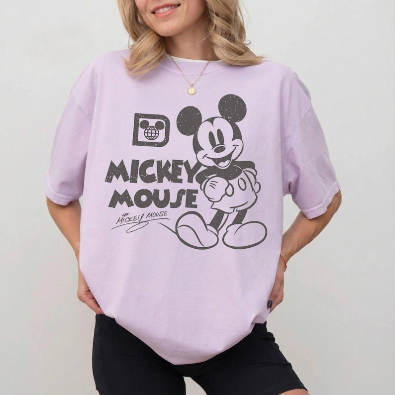May include: A light purple t-shirt featuring a distressed graphic of Mickey Mouse. The design includes the words "Mickey Mouse" in a bold font, along with Mickey's signature. The shirt has a classic crew neck and short sleeves.