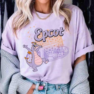 May include: A lilac-colored t-shirt featuring the word "Epcot" in a stylized font. The graphic includes a cartoon dragon character with outstretched arms, and a depiction of the Epcot ball and other park elements.
