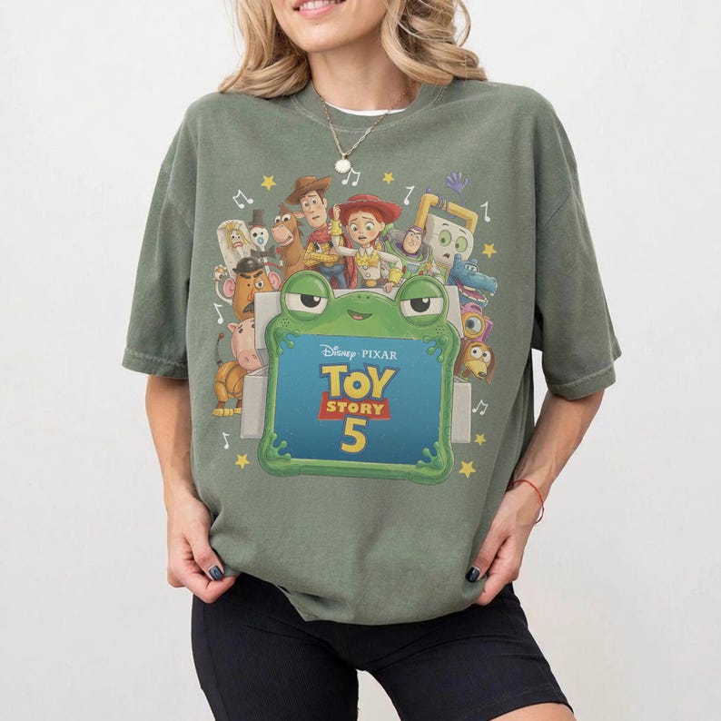 Może przedstawiać: Oliwkowozielony t-shirt z kr&oacute;tkim rękawem z nadrukiem graficznym przedstawiającym r&oacute;żne postacie z Toy Story otaczające zieloną ramkę w kształcie żaby. W środku ramki znajduje się napis Disney Pixar Toy Story 5 ż&oacute;łtymi i białymi literami.