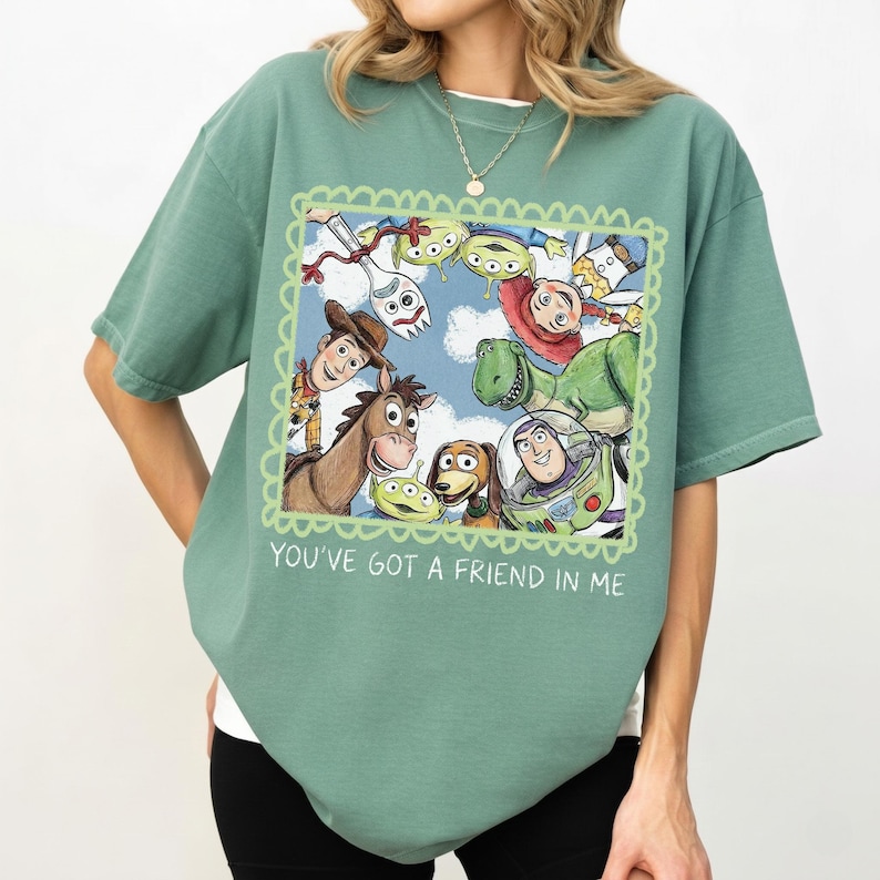 May include: Sage green t-shirt featuring a framed graphic of Toy Story characters, including Woody, Buzz Lightyear, and Rex. The graphic is surrounded by a green scalloped border, with the text "YOU'VE GOT A FRIEND IN ME" below.