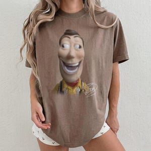 May include: A brown short-sleeve t-shirt featuring a graphic print of a smiling cowboy character with wide eyes and a yellow shirt. The character has a tan face and a red neckerchief. The word Woody is written in white cursive script below the figure.
