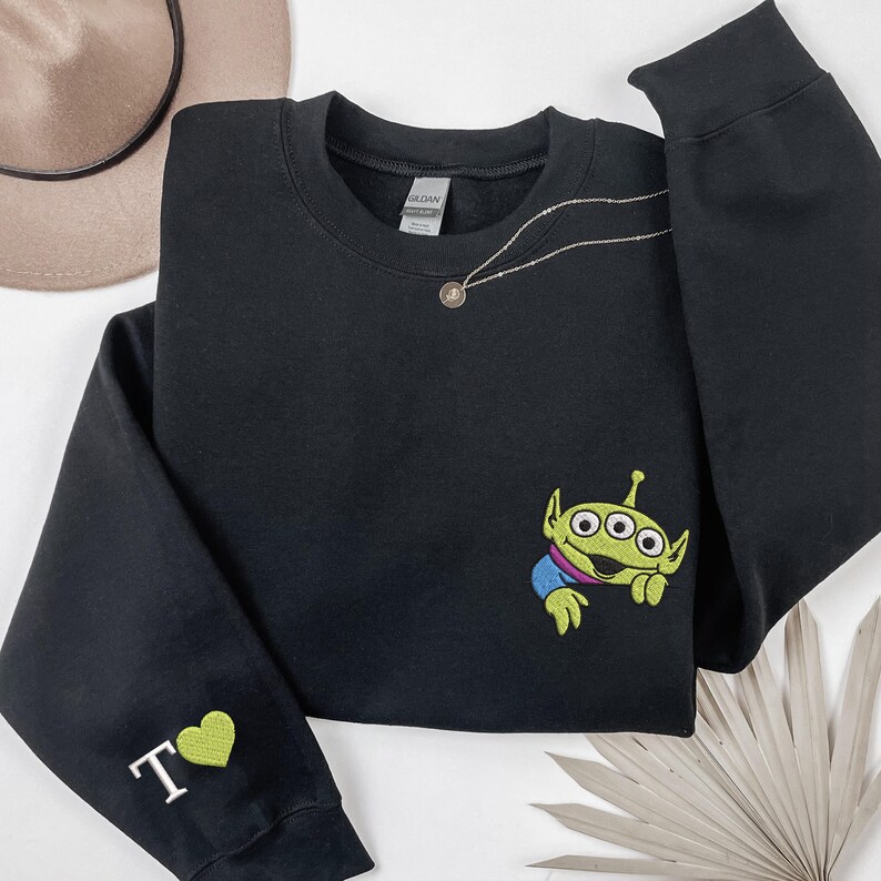 May include: A black crewneck sweatshirt featuring an embroidered green three-eyed alien character on the chest. The left sleeve displays an embroidered white letter T followed by a green heart symbol. The garment has a standard ribbed neckline.