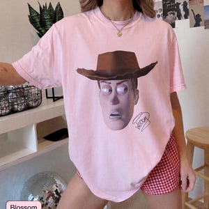 May include: A light pink short-sleeve t-shirt featuring a graphic print of the character Woody wearing a brown cowboy hat. The design includes the name Woody written in a cursive script below the character's face on the front of the garment.