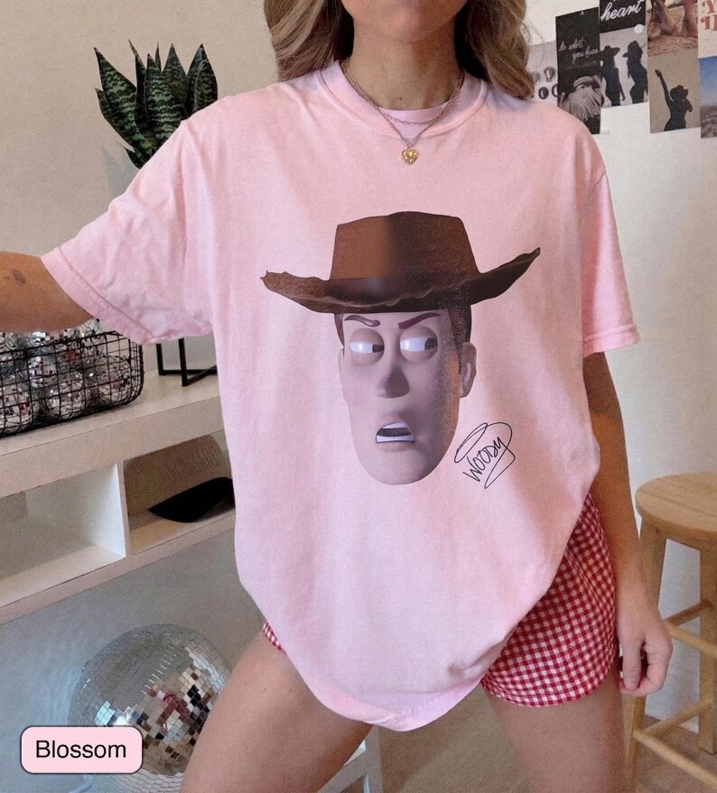 May include: A light pink short-sleeve t-shirt featuring a graphic print of the character Woody wearing a brown cowboy hat. The design includes the name Woody written in a cursive script below the character's face on the front of the garment.