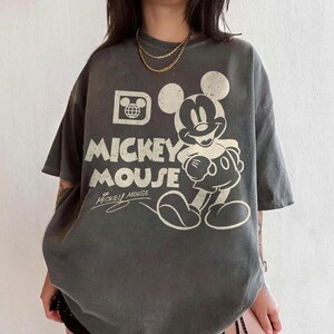 May include: Gray oversized t-shirt featuring a distressed Mickey Mouse graphic. The design includes the words "Mickey Mouse" in large, bold letters, along with a smaller signature. The shirt has short sleeves and a relaxed fit, perfect for casual wear.