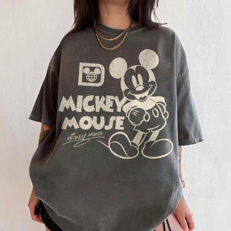 May include: Gray oversized t-shirt featuring a distressed Mickey Mouse graphic. The design includes the words "Mickey Mouse" in large, bold letters, along with a smaller signature. The shirt has short sleeves and a relaxed fit, perfect for casual wear.