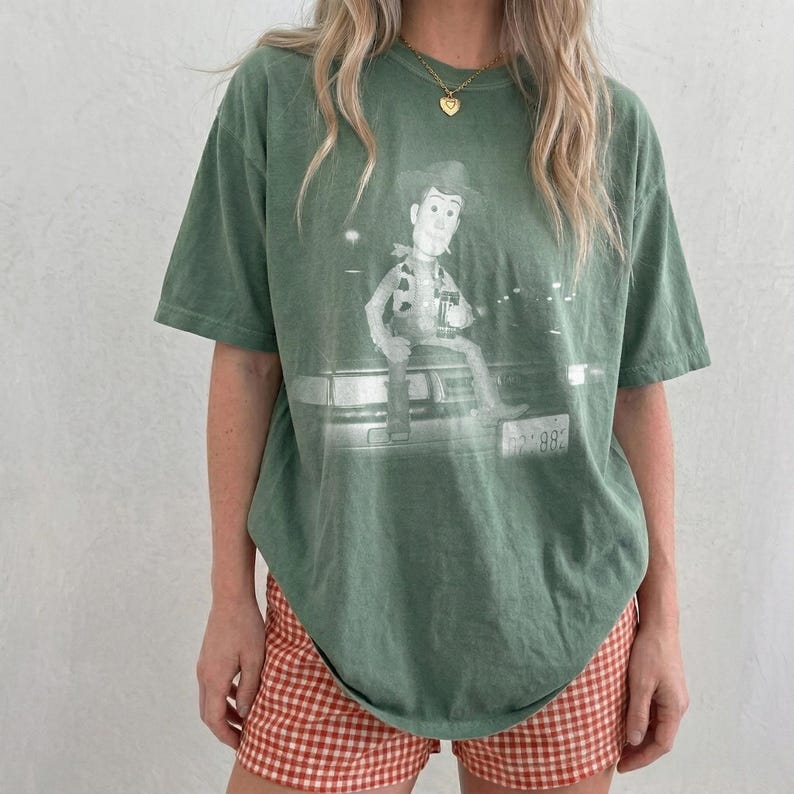 May include: A sage green short-sleeve t-shirt featuring a graphic print of the character Woody from Toy Story sitting on the hood of a car. The graphic is rendered in a faded white tone, and the shirt has a crew neckline and a relaxed fit.