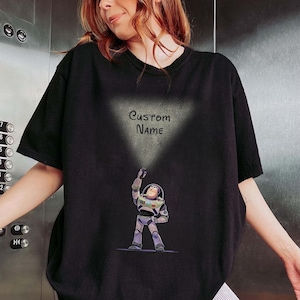 Pu&ograve; includere: T-shirt nero con un'illustrazione a cartoni animati di Buzz Lightyear di Toy Story. Il personaggio tiene in mano una torcia, con la scritta "Custom Name" sopra. Il disegno &egrave; stampato su una maglietta nera a maniche corte.