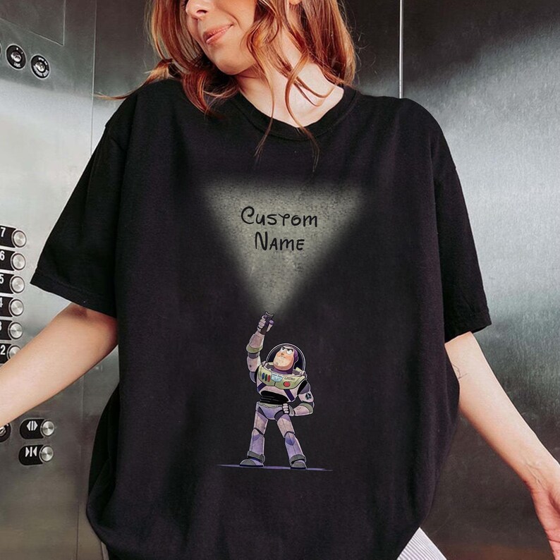 Pu&ograve; includere: T-shirt nero con un'illustrazione a cartoni animati di Buzz Lightyear di Toy Story. Il personaggio tiene in mano una torcia, con la scritta "Custom Name" sopra. Il disegno &egrave; stampato su una maglietta nera a maniche corte.