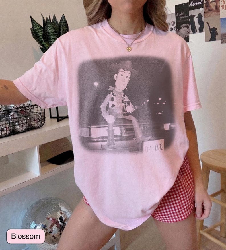 May include: A light pink short-sleeve t-shirt featuring a black and white graphic print of a cowboy character sitting on the hood of a car. The graphic includes a license plate with the text 02:882 visible at the bottom right corner.
