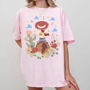 May include: A light pink t-shirt featuring a graphic illustration of a cowgirl character wearing a red hat, riding a brown horse. The design includes a cactus, various colorful flowers, blue clouds, yellow stars, and black musical notes.