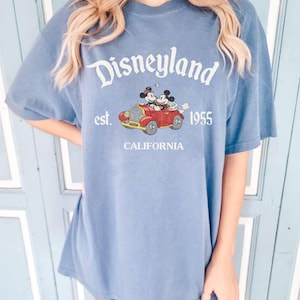K&ouml;nnte beinhalten: Blaues T-Shirt mit Disneyland-Logo, Mickey und Minnie Mouse in einem roten Auto und dem Text "est. 1955 California". Das Shirt hat kurze &Auml;rmel und eine lockere Passform.