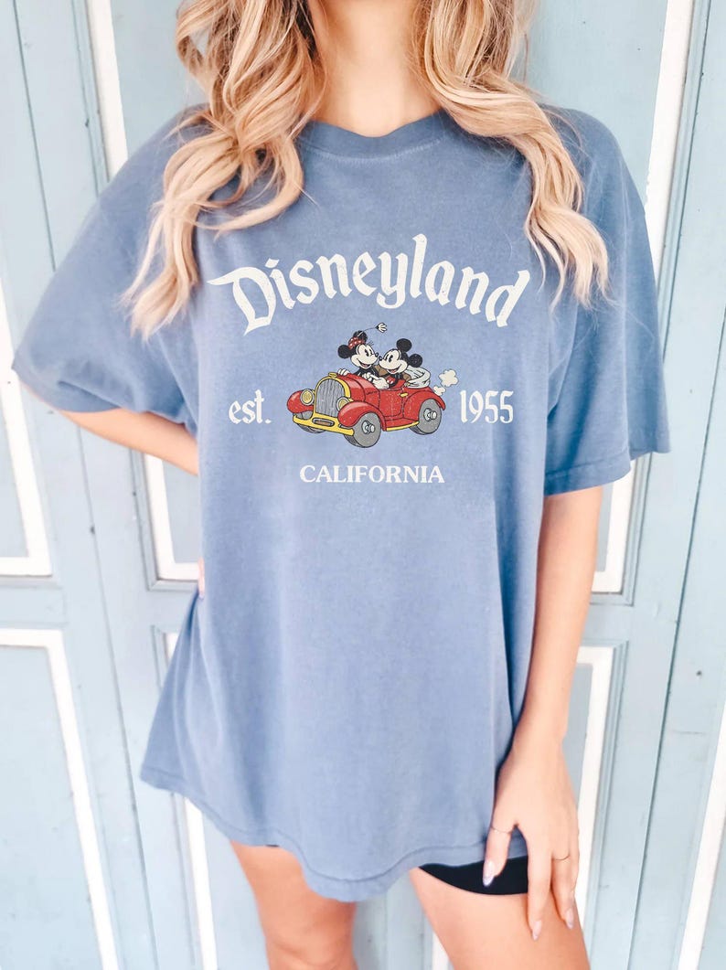K&ouml;nnte beinhalten: Blaues T-Shirt mit Disneyland-Logo, Mickey und Minnie Mouse in einem roten Auto und dem Text "est. 1955 California". Das Shirt hat kurze &Auml;rmel und eine lockere Passform.