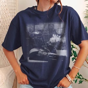 May include: A navy blue short-sleeve t-shirt featuring a monochromatic graphic print of a helmeted character in armor sitting in a tiled room, holding a smoking object. The design shows the character in a relaxed pose against a grid-like wall.