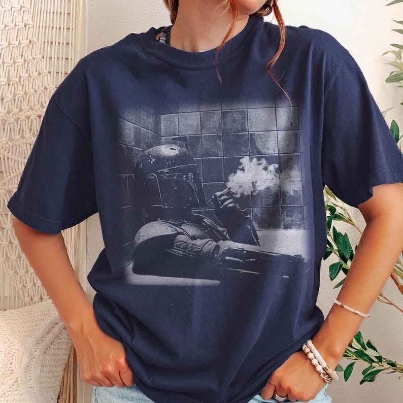 May include: A navy blue short-sleeve t-shirt featuring a monochromatic graphic print of a helmeted character in armor sitting in a tiled room, holding a smoking object. The design shows the character in a relaxed pose against a grid-like wall.