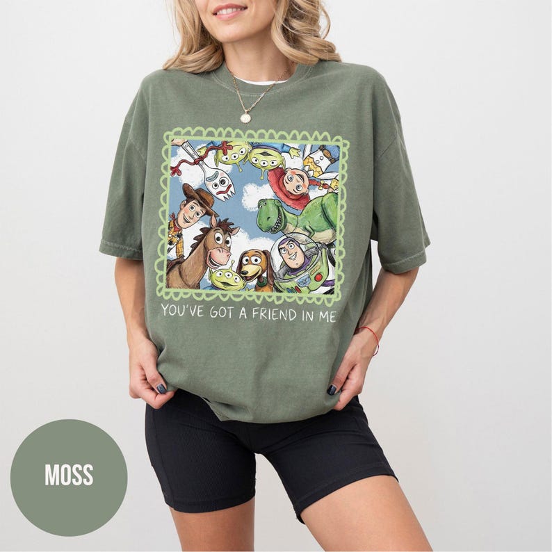 May include: Moss green t-shirt featuring a colorful graphic of characters from the animated film Toy Story. The graphic is framed with a green scalloped border and includes the text "You've Got a Friend in Me". The shirt is paired with black shorts.