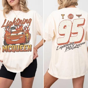 May include: Cream-colored t-shirt featuring Lightning McQueen from the Cars movie. The front graphic shows McQueen with flames and the text "Lightning McQueen". The back displays the number "95" and "Lightning McQueen".