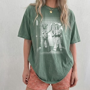 May include: A sage green short-sleeve t-shirt featuring a white graphic print of two toy characters standing side by side in front of a tiled wall. The shirt has a crew neckline and a relaxed fit, showcasing a detailed illustration of the figures.