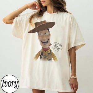 May include: An ivory short-sleeved t-shirt featuring a graphic print of the character Woody from Toy Story, wearing a brown cowboy hat, a yellow plaid shirt, and a cow-print vest. The name Woody is written in black cursive script next to the character.