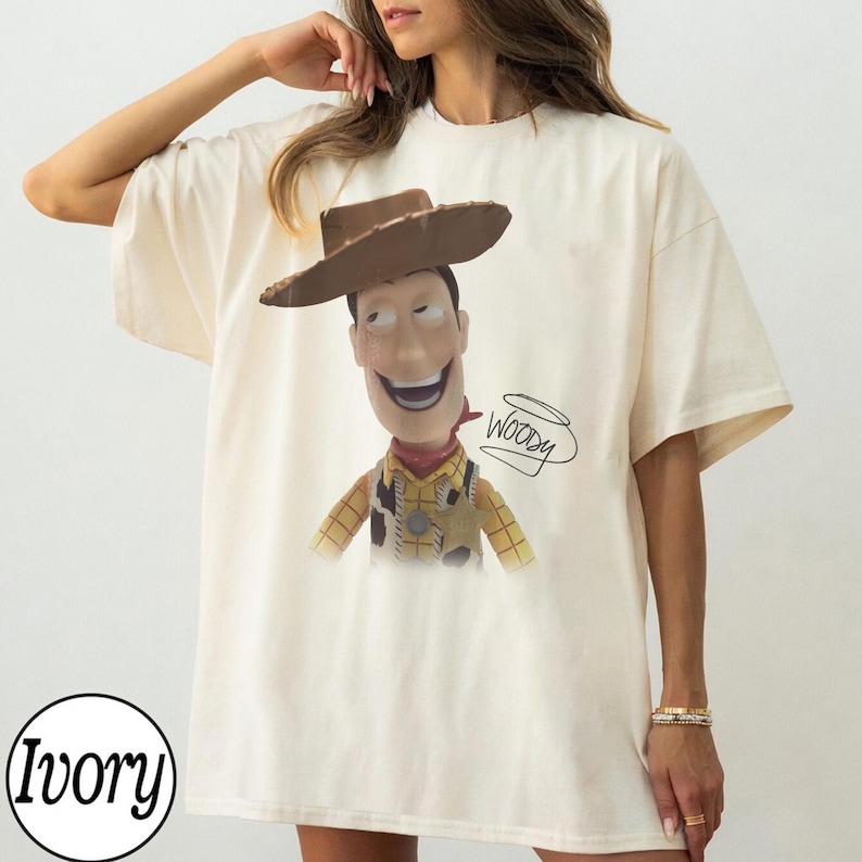May include: An ivory short-sleeved t-shirt featuring a graphic print of the character Woody from Toy Story, wearing a brown cowboy hat, a yellow plaid shirt, and a cow-print vest. The name Woody is written in black cursive script next to the character.