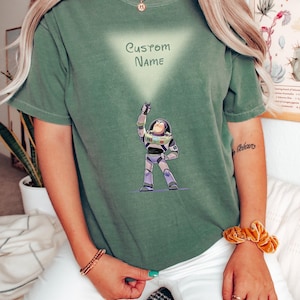 Pu&ograve; includere: T-shirt verde con un disegno a cartoni animati di Buzz Lightyear di Toy Story. Il design include il testo "CUSTOM NAME" all'interno di un triangolo chiaro. Il personaggio &egrave; raffigurato nella sua tuta spaziale, con il braccio alzato.