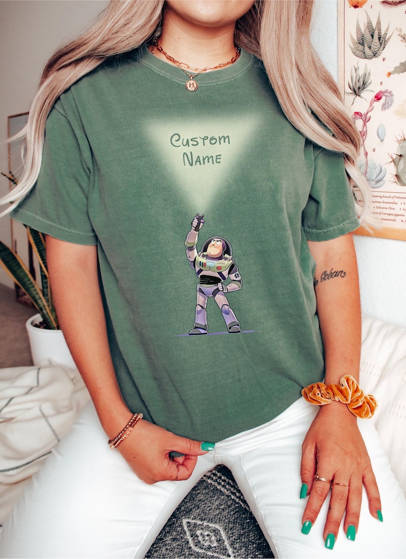 Pu&ograve; includere: T-shirt verde con un disegno a cartoni animati di Buzz Lightyear di Toy Story. Il design include il testo "CUSTOM NAME" all'interno di un triangolo chiaro. Il personaggio &egrave; raffigurato nella sua tuta spaziale, con il braccio alzato.