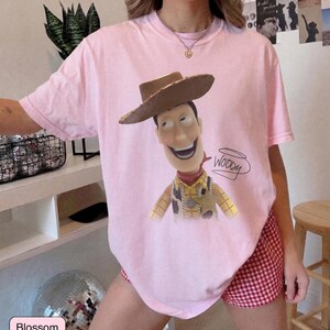 May include: A light pink short-sleeve t-shirt featuring a graphic print of the character Woody from Toy Story wearing a brown cowboy hat, a yellow plaid shirt, and a sheriff badge. The name Woody is written in black cursive script next to the character.