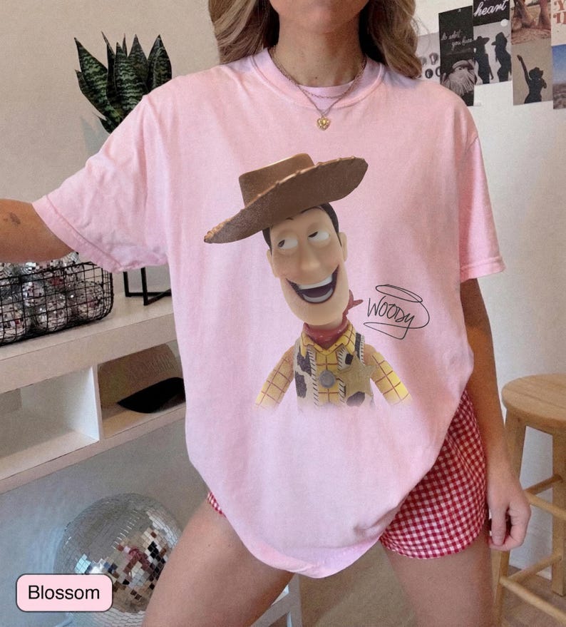 May include: A light pink short-sleeve t-shirt featuring a graphic print of the character Woody from Toy Story wearing a brown cowboy hat, a yellow plaid shirt, and a sheriff badge. The name Woody is written in black cursive script next to the character.