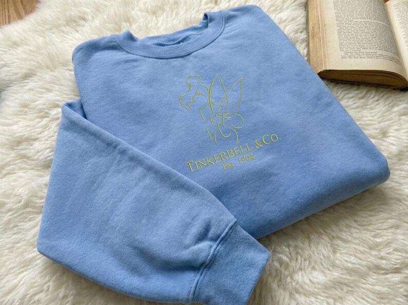 Pode incluir: Camisola azul clara com um desenho verde claro da Sininho e o texto "Tinkerbell & Co. Est. 1900". A camisola tem gola redonda e mangas compridas. &Eacute; exibida sobre uma superf&iacute;cie branca e fofa. O desenho &eacute; bordado.
