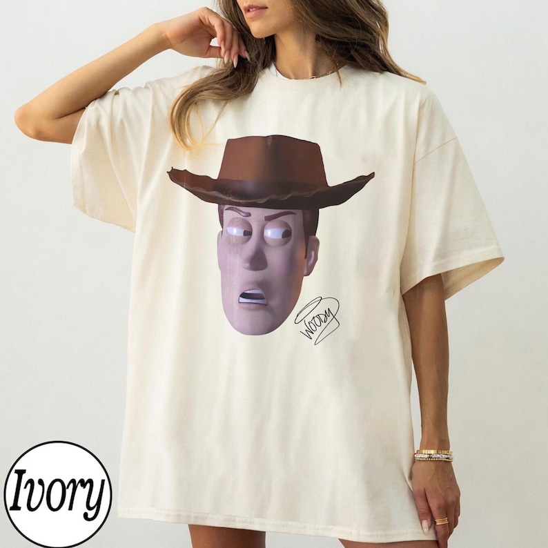 May include: An ivory t-shirt featuring a graphic print of a character wearing a brown cowboy hat. The character has a light purple complexion and a surprised expression. The word Woody is written in a cursive script below the character's chin.
