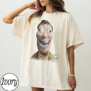 May include: An ivory short-sleeved t-shirt featuring a graphic print of a stylized, wide-eyed character head with a broad, toothy grin. The character wears a yellow collared shirt, and the word Woody is written in a cursive script below the image.