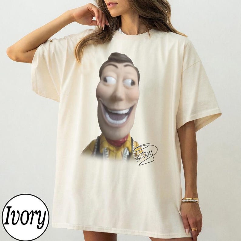 May include: An ivory short-sleeved t-shirt featuring a graphic print of a stylized, wide-eyed character head with a broad, toothy grin. The character wears a yellow collared shirt, and the word Woody is written in a cursive script below the image.